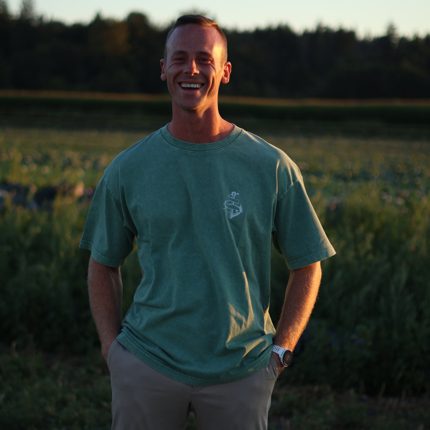 Man wearing an olive green AfterDark Project “Be Your Anchor” t-shirt with chest logo, standing outdoors in a sunlit field — designed by a Canadian streetwear brand.