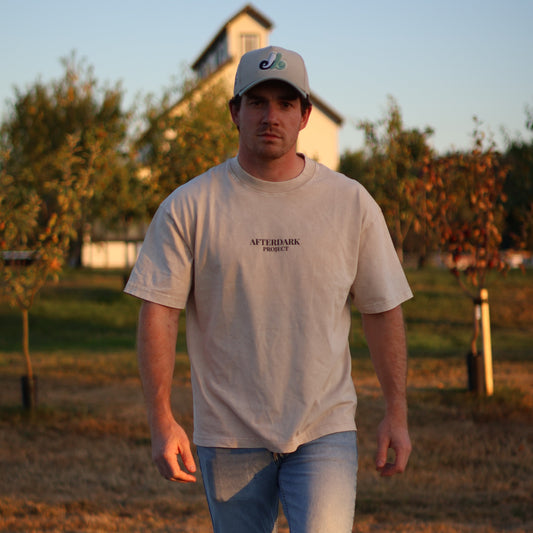 Man wearing an AfterDark Project oversized beige t-shirt and baseball cap, walking outdoors in a rustic fall setting with trees and warm sunset lighting — streetwear designed by a Canadian brand.