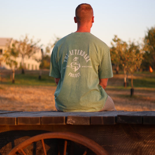 Man sitting on a rustic wooden wagon wearing an olive green AfterDark Project “Be Your Anchor” t-shirt — bold back print featuring anchor graphic, captured outdoors during golden hour in a fall landscape.