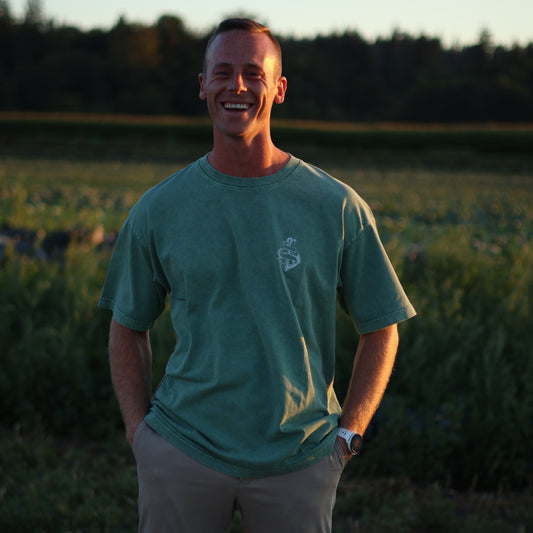 Man wearing an olive green AfterDark Project “Be Your Anchor” t-shirt with chest logo, standing outdoors in a sunlit field — designed by a Canadian streetwear brand.