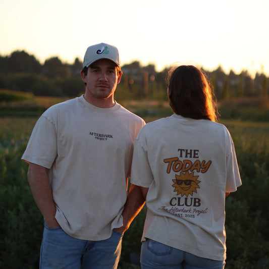 Man and woman wearing AfterDark Project “The Today Club” graphic t-shirts standing in a scenic outdoor field during golden hour — modern unisex Canadian streetwear for everyday wear.